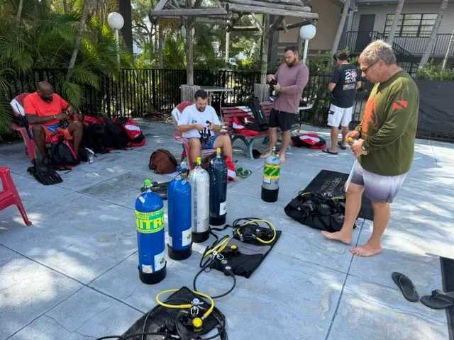 Scuba tanks and regulators laid out on a pool deck while divers get their gear ready