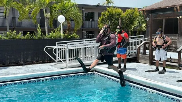 A diver in full scuba gear steps off the pool edge into the water