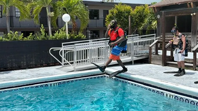 A diver in full scuba gear steps off the pool edge into the water
