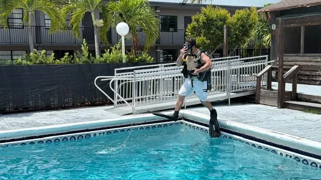 A diver in full scuba gear steps off the pool edge into the water