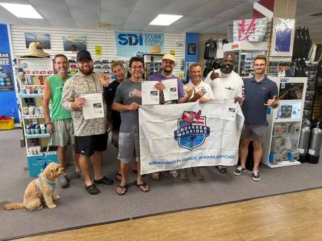 The first certified class holding certificates and the Undersea Warriors banner inside a dive shop, with the team mascot dog